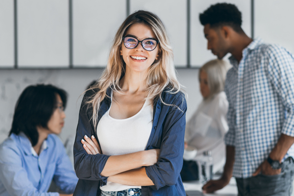 Confident Blonde Female Manager Posing With Smile After Conference With Other Employees. Asian Programmer Talking With African Freelancer While Fair Haired Secretary Laughing On Foreground..